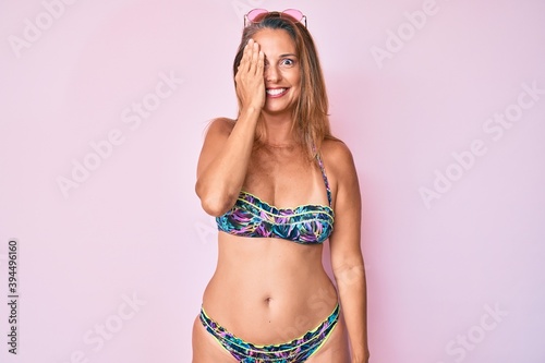 Middle age hispanic woman wearing bikini covering one eye with hand, confident smile on face and surprise emotion.