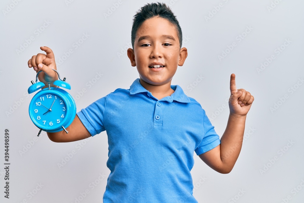 Little boy hispanic kid holding alarm clock smiling with an idea or question pointing finger with happy face, number one Little boy hispanic kid holding alarm clock smiling with an idea or question pointing finger with happy face, number one