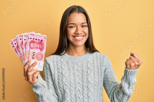 Beautiful hispanic woman holding 100 yuan chinese banknotes screaming proud, celebrating victory and success very excited with raised arm