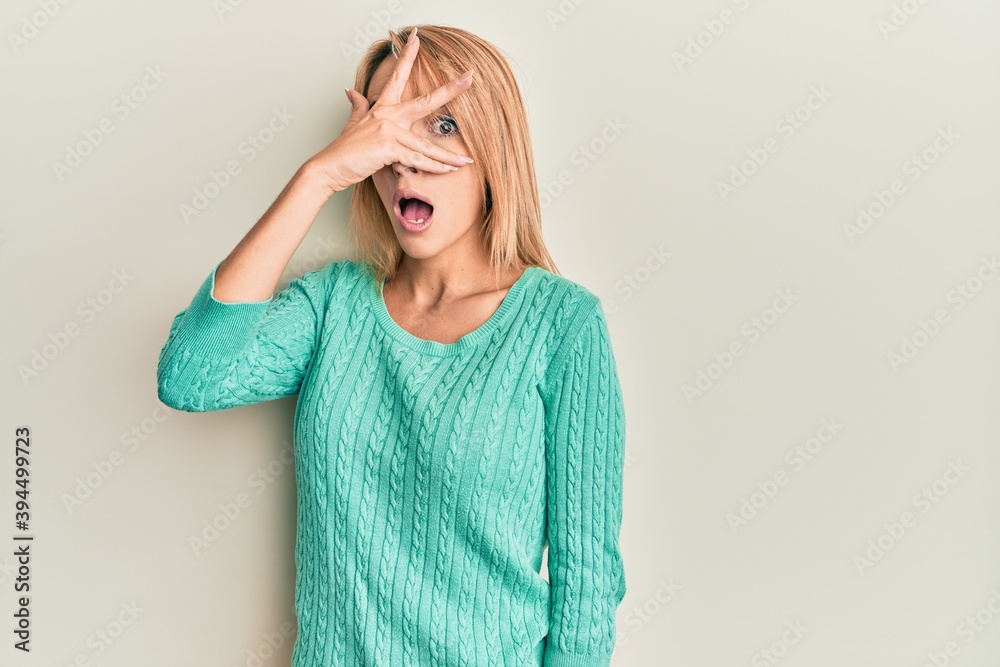Beautiful blonde woman wearing casual winter sweater peeking in shock covering face and eyes with hand, looking through fingers with embarrassed expression. Beautiful blonde woman wearing casual winter sweater peeking in shock covering face and eyes with hand, looking through fingers with embarrassed expression.