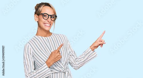 Beautiful blonde woman wearing business shirt and glasses smiling and looking at the camera pointing with two hands and fingers to the side.