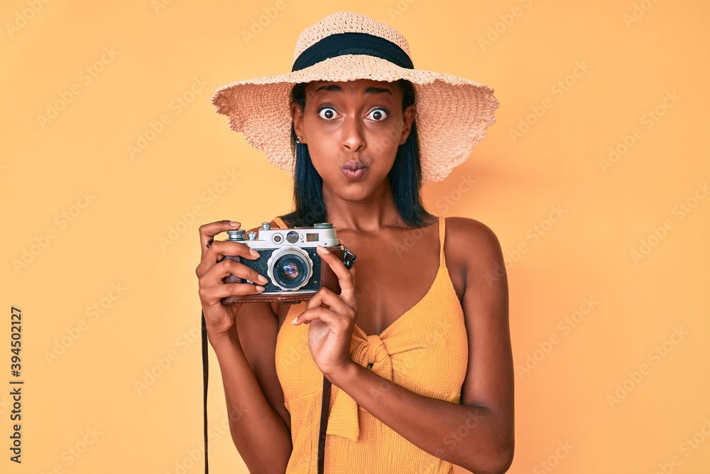 Young african american woman wearing summer hat holding vintage camera puffing cheeks with funny face. mouth inflated with air, catching air. Young african american woman wearing summer hat holding vintage camera puffing cheeks with funny face. mouth inflated with air, catching air.