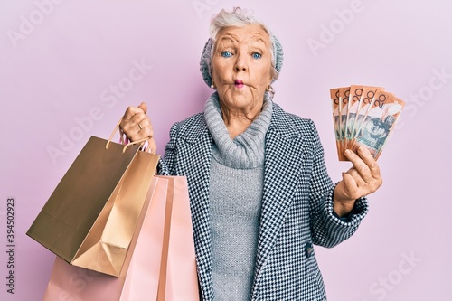 Senior grey-haired woman holding shopping bags and australia dollars making fish face with mouth and squinting eyes, crazy and comical.