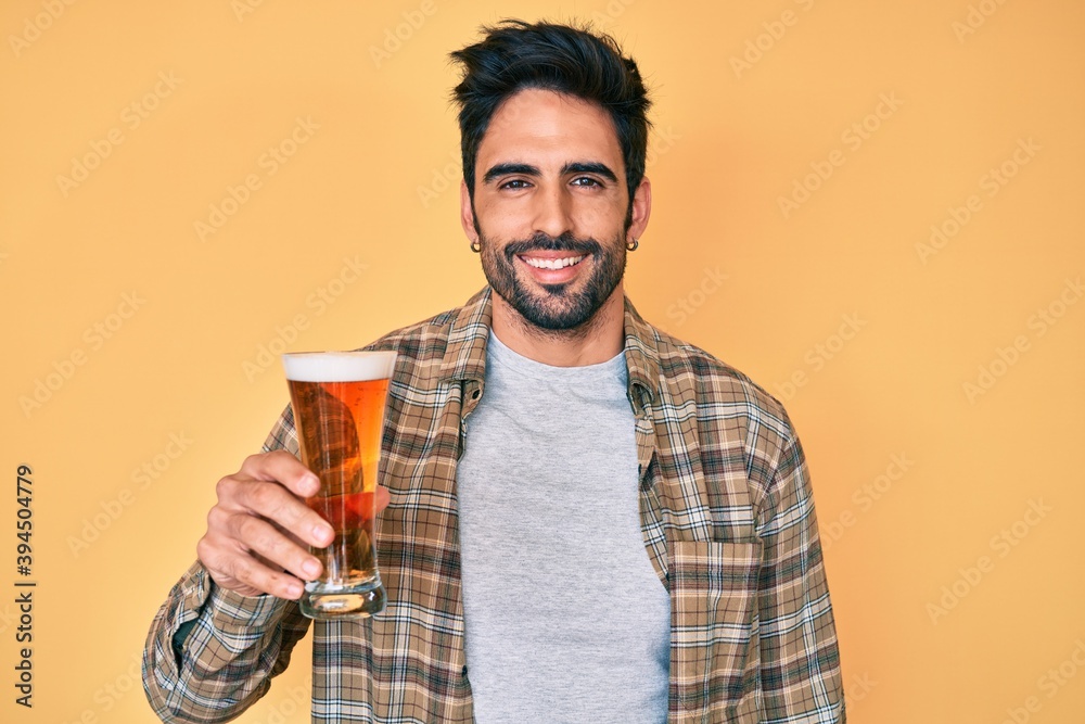 Handsome hispanic man with beard drinking a pint of beer looking positive and happy standing and smiling with a confident smile showing teeth Handsome hispanic man with beard drinking a pint of beer looking positive and happy standing and smiling with a confident smile showing teeth