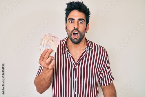 Handsome hispanic man with beard holding united kingdom pounds scared and amazed with open mouth for surprise, disbelief face