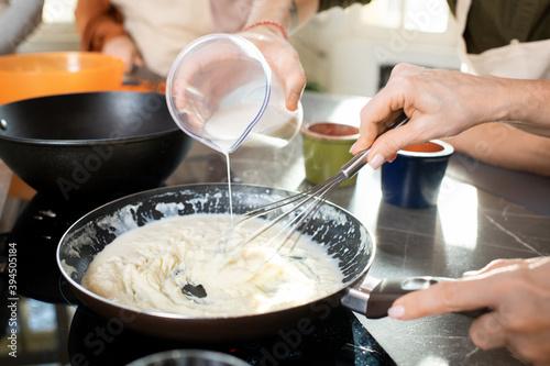 Hands of young man pouring milk into frying pan with flour while cooking
