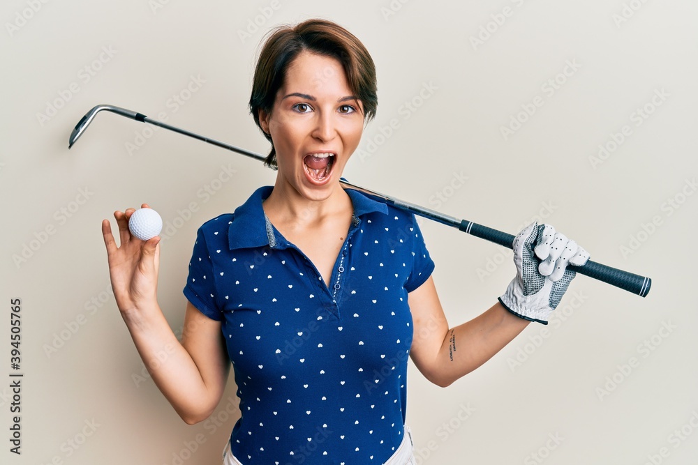 Young brunette woman with short hair holding ball and golf club celebrating crazy and amazed for success with open eyes screaming excited. Young brunette woman with short hair holding ball and golf club celebrating crazy and amazed for success with open eyes screaming excited.
