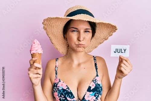 Young brunette woman with short hair holding ice cream and july paper puffing cheeks with funny face. mouth inflated with air, catching air.