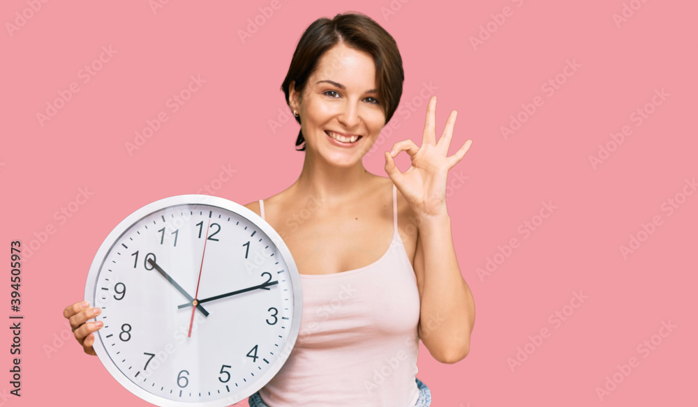 Young brunette woman with short hair holding big clock doing ok sign with fingers, smiling friendly gesturing excellent symbol Young brunette woman with short hair holding big clock doing ok sign with fingers, smiling friendly gesturing excellent symbol