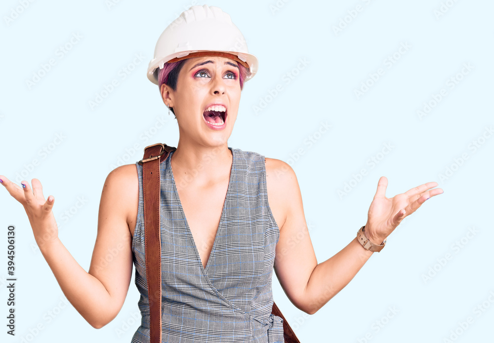 Young beautiful woman wearing architect hardhat and leather bag crazy and mad shouting and yelling with aggressive expression and arms raised. frustration concept. Young beautiful woman wearing architect hardhat and leather bag crazy and mad shouting and yelling with aggressive expression and arms raised. frustration concept.