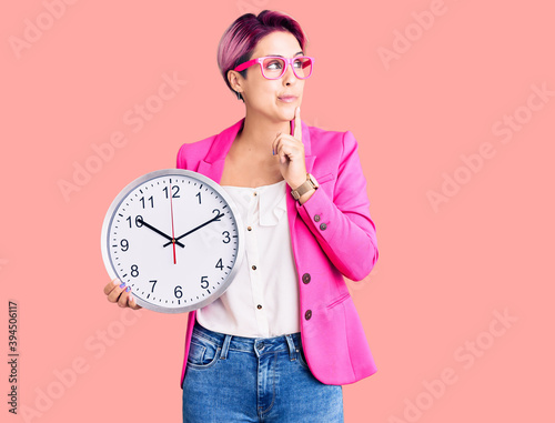 Young beautiful woman with pink hair wearing business jacket and holding clock serious face thinking about question with hand on chin, thoughtful about confusing idea