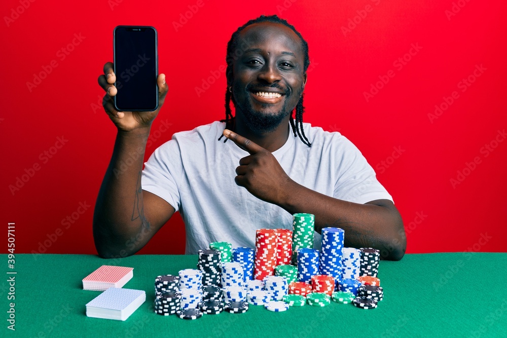 Handsome young black man playing poker holding smartphone smiling happy pointing with hand and finger