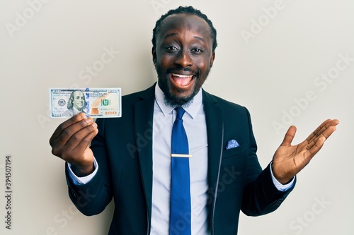 Handsome young black man wearing business suit and tie holding 100 dollars celebrating achievement with happy smile and winner expression with raised hand