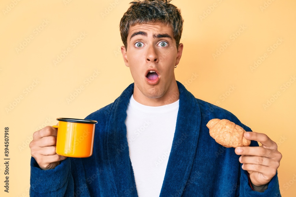 Young handsome man wearing bathrobe eating breakfast holding coffee and croissant afraid and shocked with surprise and amazed expression, fear and excited face. Young handsome man wearing bathrobe eating breakfast holding coffee and croissant afraid and shocked with surprise and amazed expression, fear and excited face.