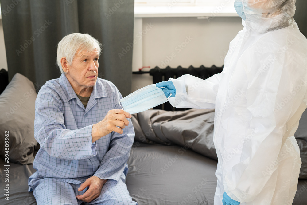 Obraz premium Sick senior man in blue pajamas taking medical mask from gloved hand of doctor