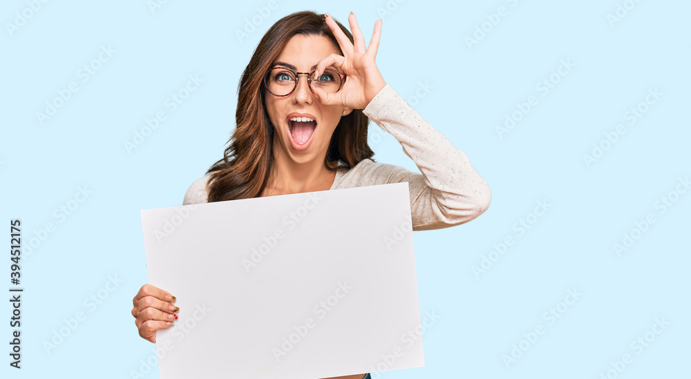 Naklejka premium Young brunette woman holding blank empty banner smiling happy doing ok sign with hand on eye looking through fingers