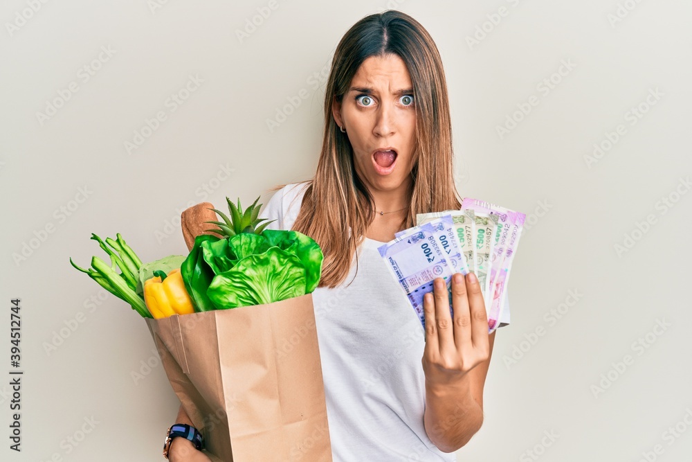 Brunette young woman holding groceries and indian rupee banknotes afraid and shocked with surprise and amazed expression, fear and excited face. Brunette young woman holding groceries and indian rupee banknotes afraid and shocked with surprise and amazed expression, fear and excited face.