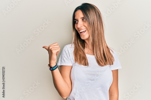 Brunette young woman wearing casual white t shirt pointing thumb up to the side smiling happy with open mouth