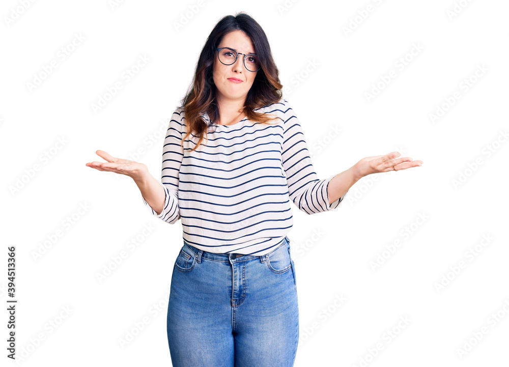 Beautiful young brunette woman wearing casual clothes and glasses clueless and confused expression with arms and hands raised. doubt concept. Beautiful young brunette woman wearing casual clothes and glasses clueless and confused expression with arms and hands raised. doubt concept.