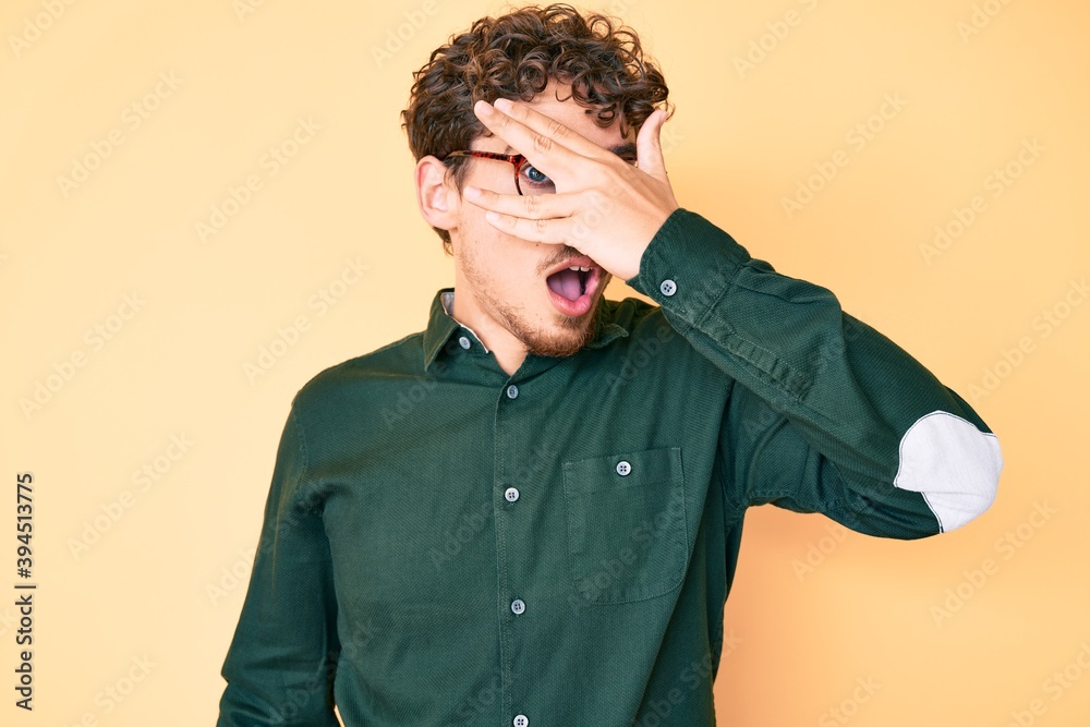 Young caucasian man with curly hair wearing casual clothes and glasses peeking in shock covering face and eyes with hand, looking through fingers with embarrassed expression. Young caucasian man with curly hair wearing casual clothes and glasses peeking in shock covering face and eyes with hand, looking through fingers with embarrassed expression.