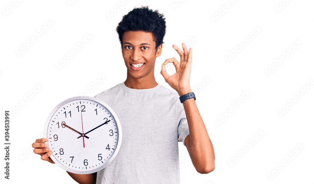 Young african american man holding big clock doing ok sign with fingers, smiling friendly gesturing excellent symbol Young african american man holding big clock doing ok sign with fingers, smiling friendly gesturing excellent symbol