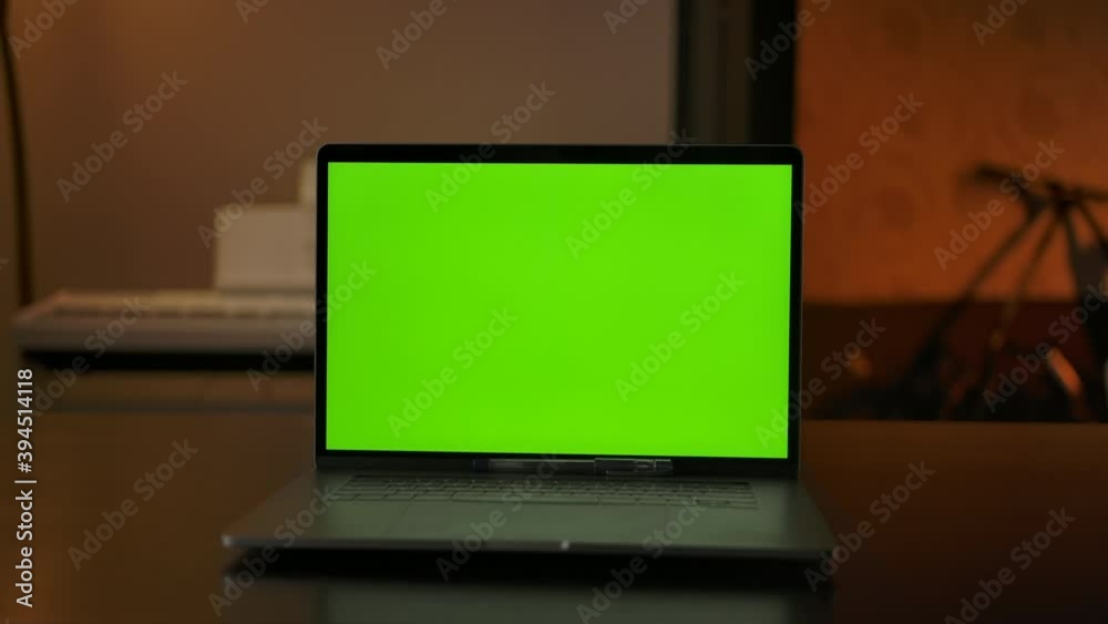 Laptop computer showing green chroma key screen stands on a desk in the living room. Cozy living room in the evening with warm lights on In the background. Dolly tracking left to right shot. Laptop computer showing green chroma key screen stands on a desk in the living room. Cozy living room in the evening with warm lights on In the background. Dolly tracking left to right shot.