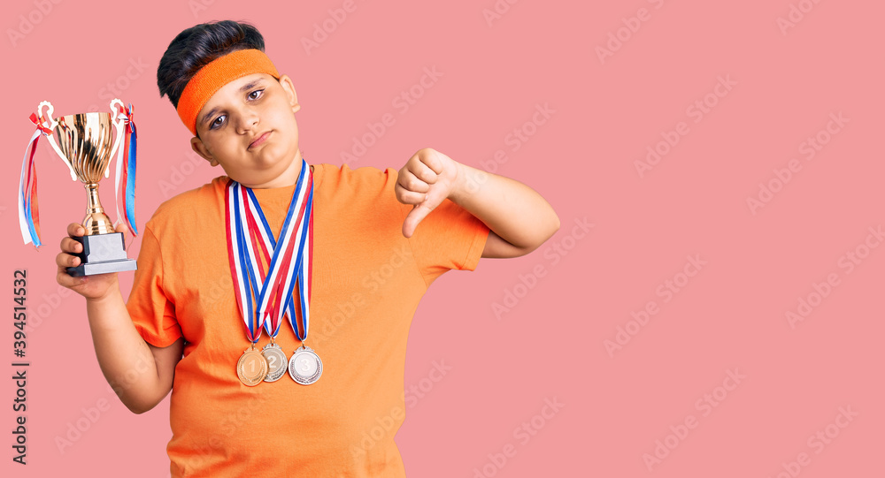 Little boy kid holding champion trophy and wearing medals with angry face, negative sign showing dislike with thumbs down, rejection concept Little boy kid holding champion trophy and wearing medals with angry face, negative sign showing dislike with thumbs down, rejection concept