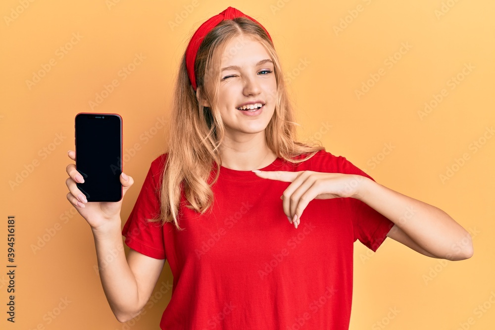 Naklejka premium Beautiful young caucasian girl holding smartphone showing screen winking looking at the camera with sexy expression, cheerful and happy face.