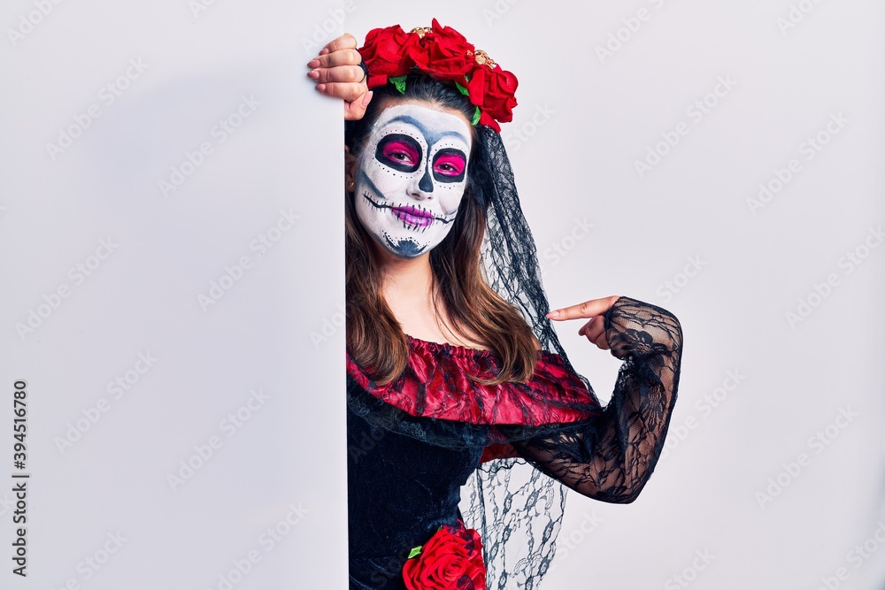 Young woman wearing day of the dead custome holding blank empty banner looking confident with smile on face, pointing oneself with fingers proud and happy. Young woman wearing day of the dead custome holding blank empty banner looking confident with smile on face, pointing oneself with fingers proud and happy.