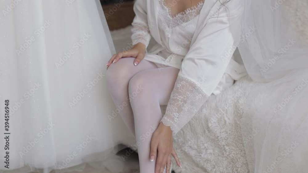 Slim legs of young bride wearing stylish white lacy rest gown and high heeled shoes sitting on bed in bedroom. Woman put on wearing wedding shoes. Bridal footwear, morning dressing before ceremony Slim legs of young bride wearing stylish white lacy rest gown and high heeled shoes sitting on bed in bedroom. Woman put on wearing wedding shoes. Bridal footwear, morning dressing before ceremony