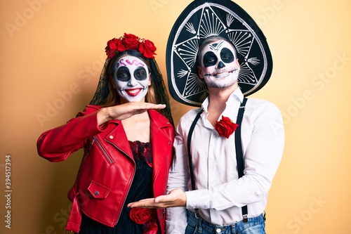 Couple wearing day of the dead costume over yellow gesturing with hands showing big and large size sign, measure symbol. smiling looking at the camera. measuring concept.
