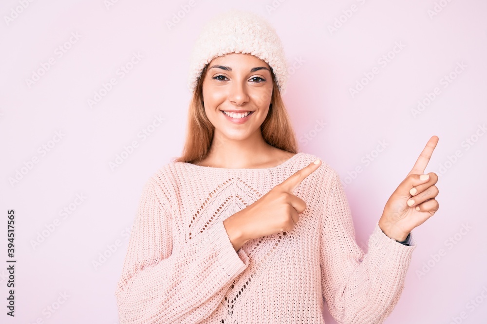 Young brunette woman wearing winter hat and sweater smiling and looking at the camera pointing with two hands and fingers to the side. Young brunette woman wearing winter hat and sweater smiling and looking at the camera pointing with two hands and fingers to the side.