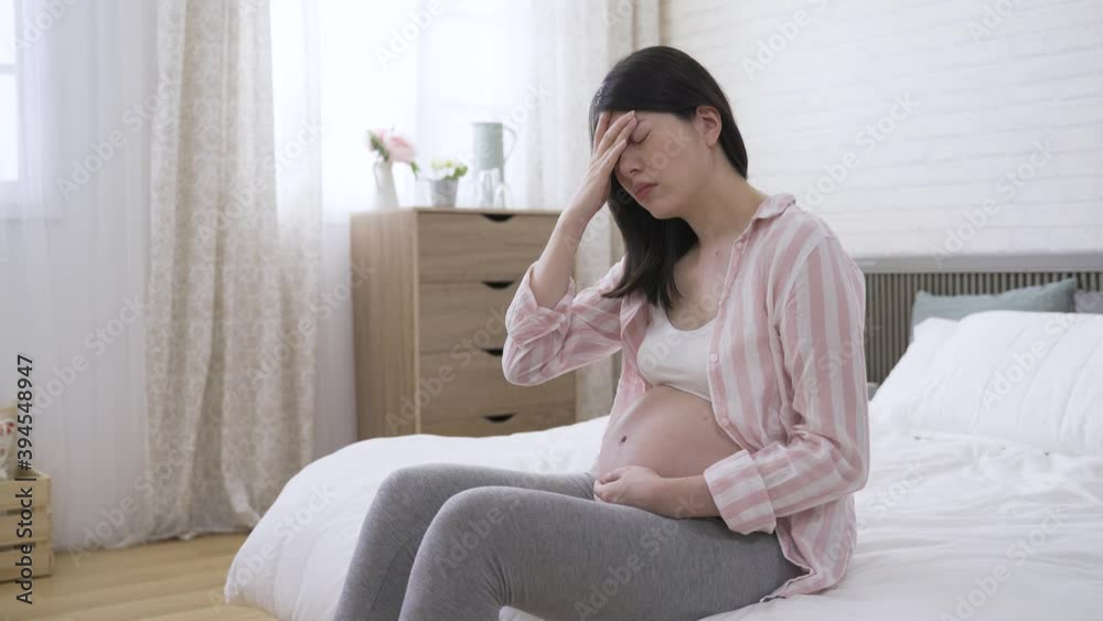 chinese pregnant lady sitting on bed and touching her forehead with painful look is suffering dizziness at bright home interior. chinese pregnant lady sitting on bed and touching her forehead with painful look is suffering dizziness at bright home interior.