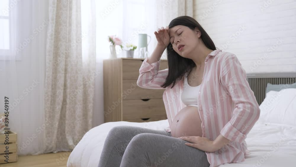 sweaty pregnant lady seated on bed at home is mopping her forehead and