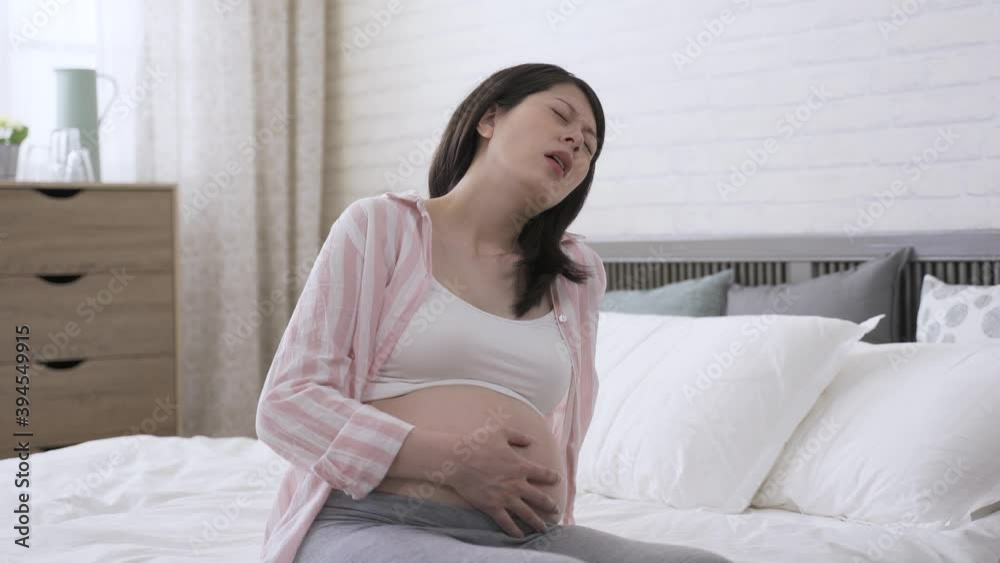 asian expectant mother suffering labor contraction is moaning and holding her tummy while her face is contorted in a grimace of pain in the bedroom. asian expectant mother suffering labor contraction is moaning and holding her tummy while her face is contorted in a grimace of pain in the bedroom.