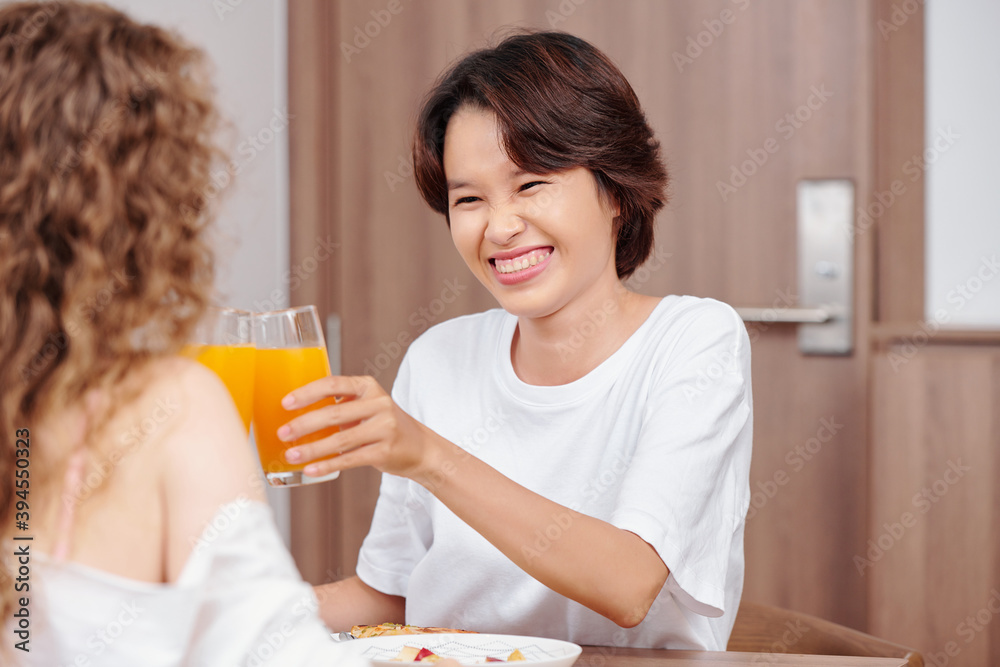Cheerful pretty young Vietnamse woman toasting with glass of juice when eating breakfast with girlfriend Cheerful pretty young Vietnamse woman toasting with glass of juice when eating breakfast with girlfriend