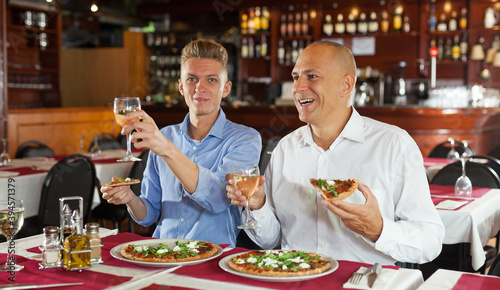 Two happy men emotionally watching sports on tv while eating pizza with white wine in cozy restaurant