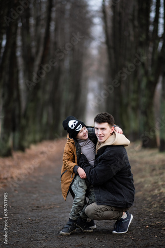 father and son are walking in the park, sitting in the middle of the alley, single father, cold spring, autumn, winter, against the background of large trees, have fun smiling, boy sits on his knees © Maxim