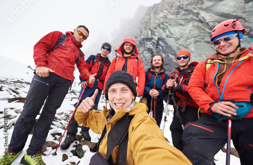 Young man taking picture with friends hikers during travel in winter mountains. Group of male travelers looking at camera and smiling, showing thumbs up. Concept of travelling, hiking and photography.