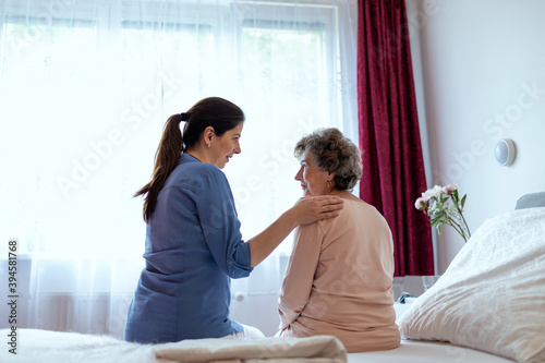 Home Caregiver Comforting Senior Woman Patient Sitting in Her Bedroom. Nurse Putting Her Hand Over Elderly Patient Shoulder in Hospital Room.