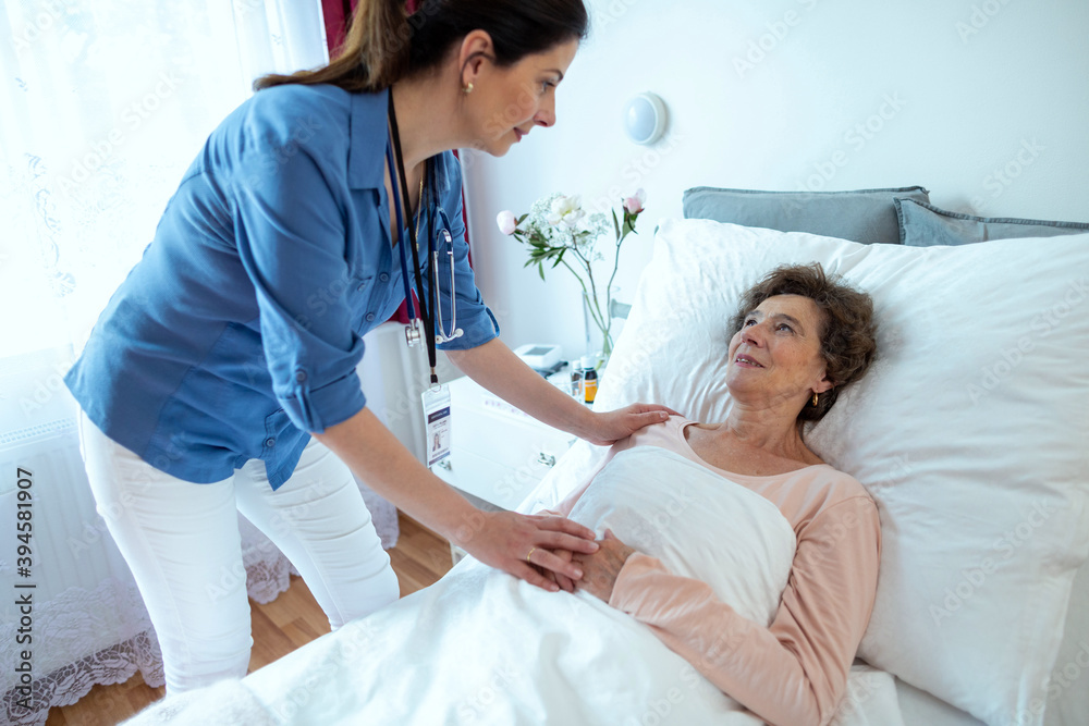 Fototapeta premium Nurse Leaning Over Elderly Woman Lying in Hospital Bed. Senior Female Patient Looking at Home Caregiver Holding Her Hand.