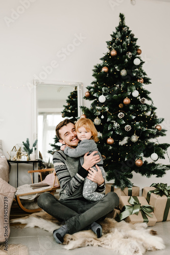 Father and little child having fun and playing together at home. Portrait loving family close up. Cheerful dad hugging cute baby daughter girl near Christmas tree. Merry Christmas and Happy Holidays. © Serhii