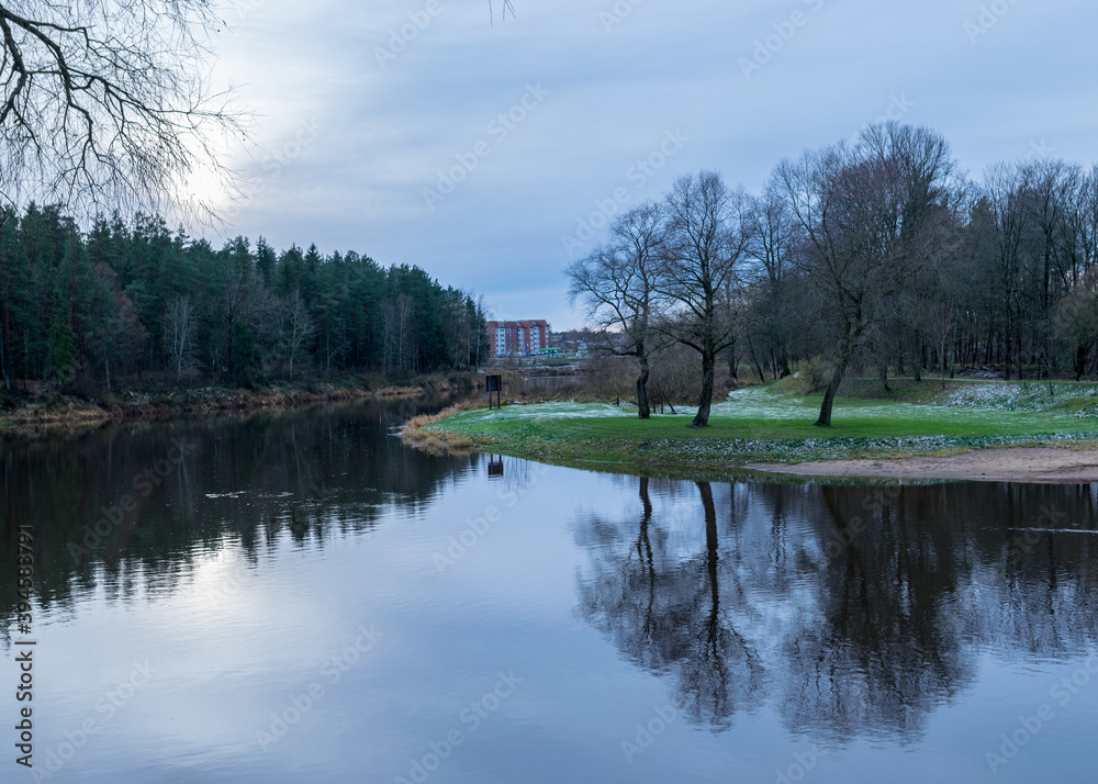 dark and bare tree silhouettes reflect in dark river water, late autumn, first snow on green grass, dark and bare tree silhouettes reflect in dark river water, late autumn, first snow on green grass,