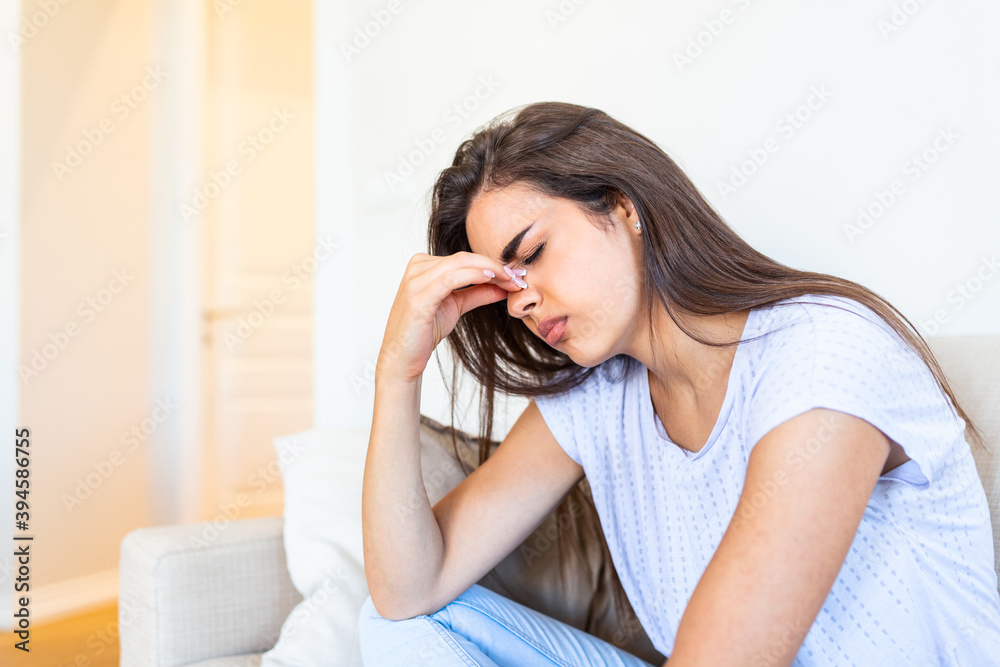 Portrait of an attractive woman sitting on a sofa at home with a headache, feeling pain and with an expression of being unwell. Upset depressed woman sitting on couch feeling strong headache migraine. Portrait of an attractive woman sitting on a sofa at home with a headache, feeling pain and with an expression of being unwell. Upset depressed woman sitting on couch feeling strong headache migraine.