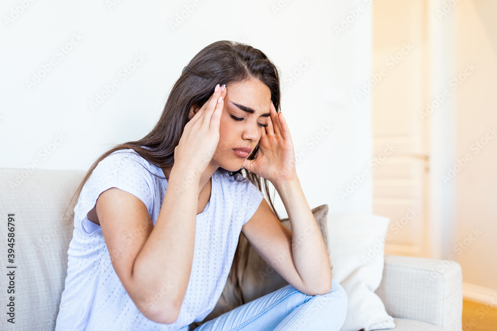Portrait of an attractive woman sitting on a sofa at home with a headache, feeling pain and with an expression of being unwell. Upset woman sitting on couch feeling strong headache migraine. Portrait of an attractive woman sitting on a sofa at home with a headache, feeling pain and with an expression of being unwell. Upset woman sitting on couch feeling strong headache migraine.