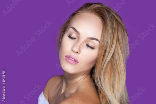 Portrait of a sexy blonde young woman with makeup and blong hair with bare shoulders, over bright purple background.