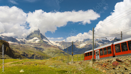 Postcard photo of the Matterhorn and the red train heading towards it with a clear sky and at the same time some clouds