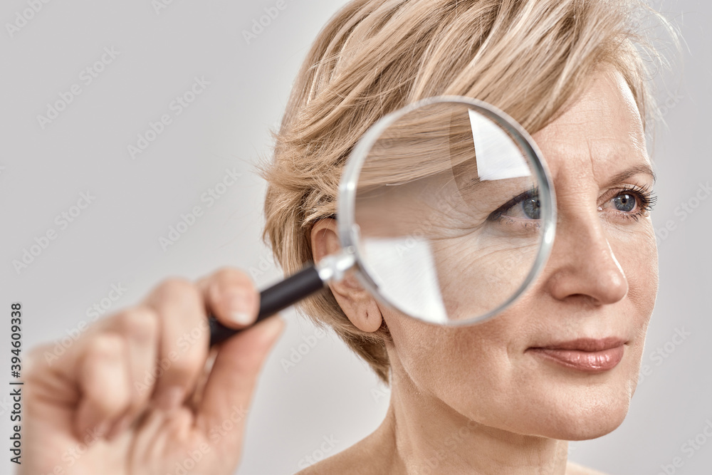 Close up portrait of attractive middle aged woman looking aside, holding a magnifying glass and showing her wrinkles, posing isolated over grey background Close up portrait of attractive middle aged woman looking aside, holding a magnifying glass and showing her wrinkles, posing isolated over grey background