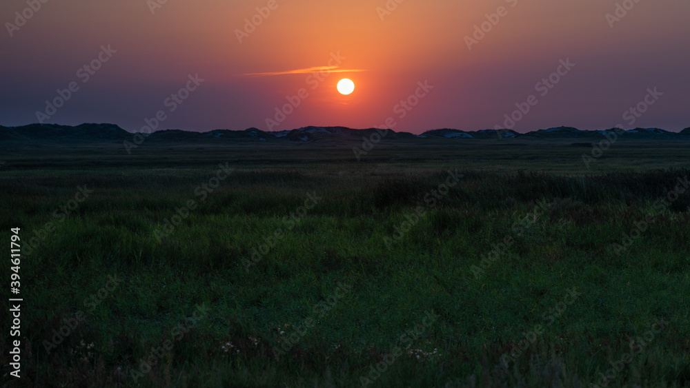 Panoramic view over the grassland between the dike and the beach against the deep setting sun on the North Sea coast.