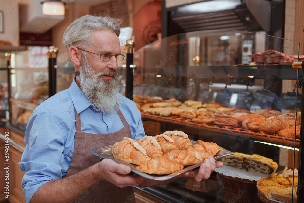Cheerfyl elderly male baker carrying croissants to bake at his bakery store Cheerfyl elderly male baker carrying croissants to bake at his bakery store